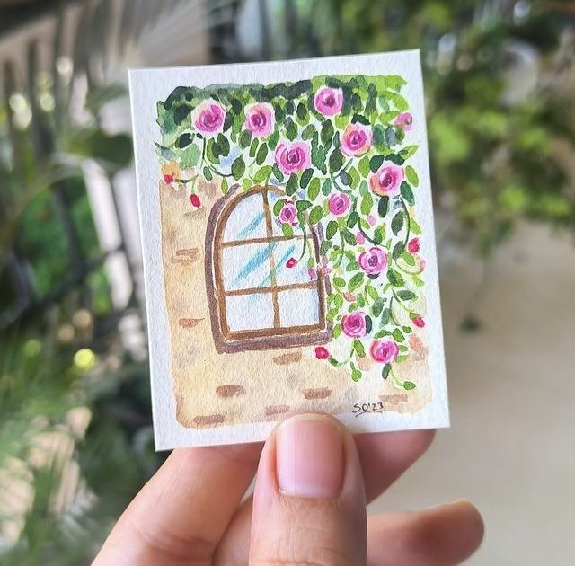 a hand holding up a small card with a window and flowers on the outside wall