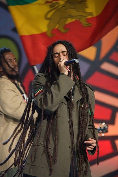 two men with dreadlocks singing into microphones in front of a large flag