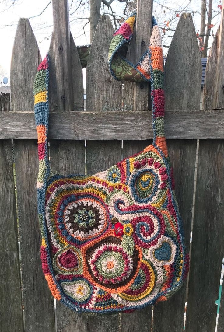 a multicolored crocheted purse hanging on a wooden fence with a tree in the background