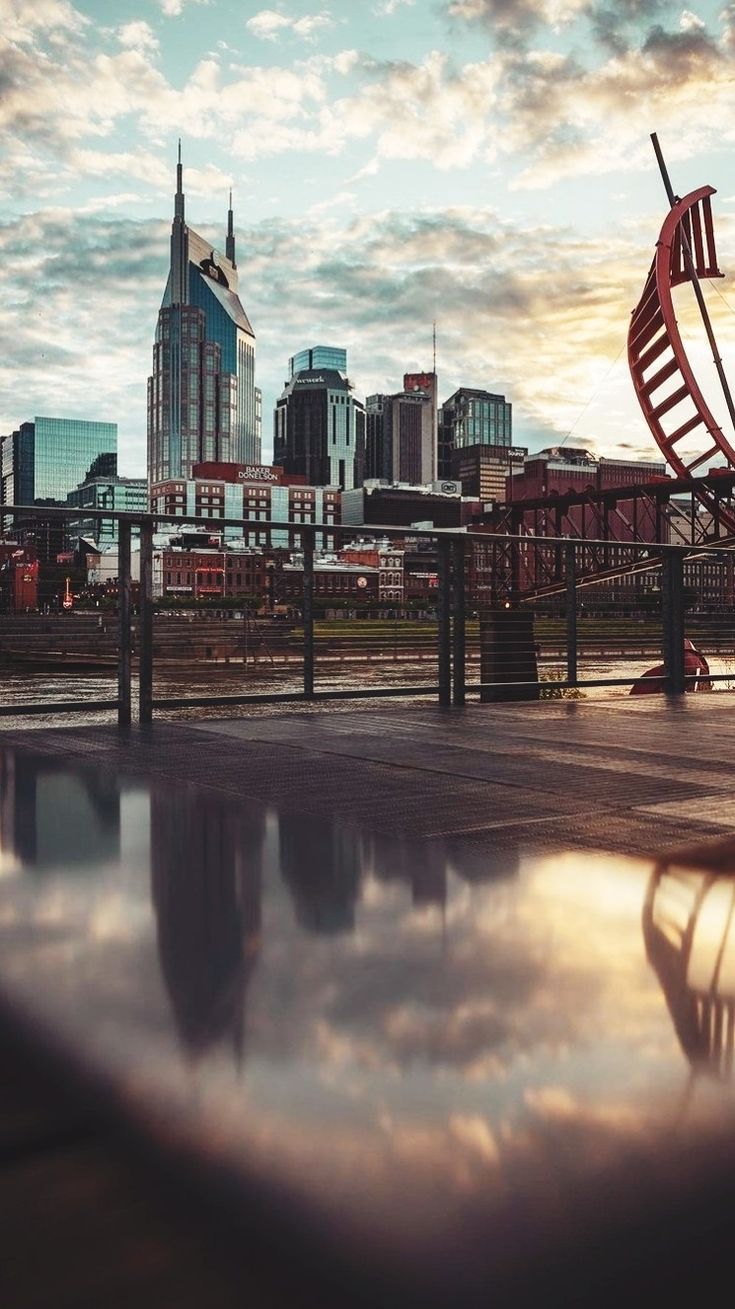 a city skyline is reflected in a puddle on the ground as it sits behind a metal fence