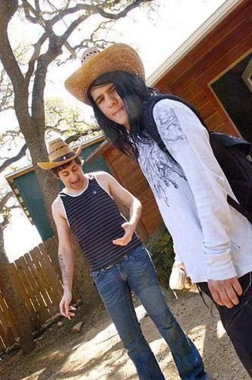 two young men standing next to each other in front of a house and trees with one man wearing a cowboy hat