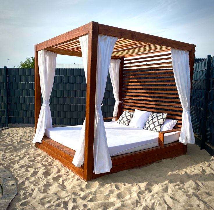 a wooden bed with white drapes and pillows on the sand in front of a fence