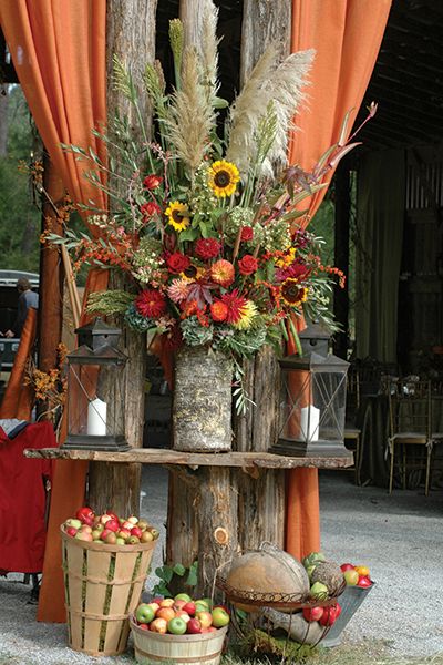 an arrangement of flowers and fruit is displayed in front of a wooden structure with orange drapes