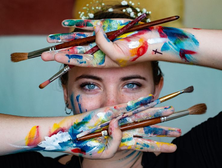 a woman holding her hands up to her face with paint on it and brushes in front of her