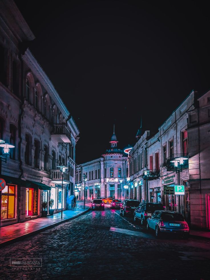 an empty city street at night with cars parked on the side and buildings lit up