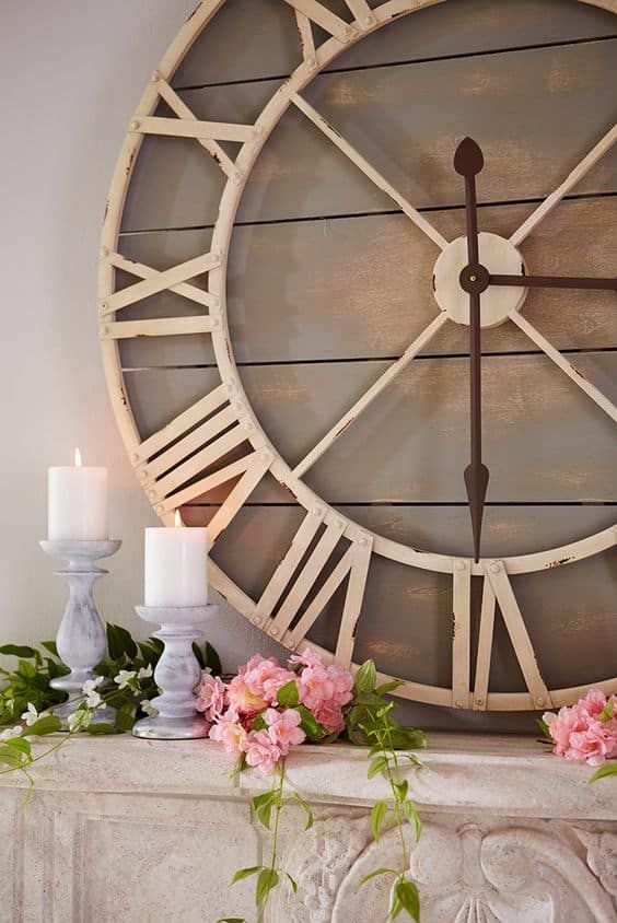 a large clock sitting on top of a mantle next to flowers and two white candles