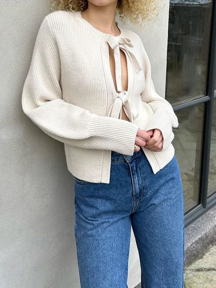 a woman leaning against a wall with her hands on her hips wearing jeans and a sweater