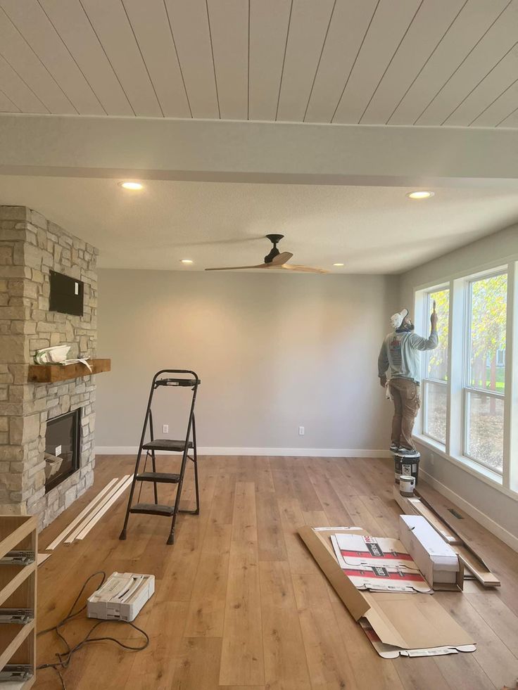 a man standing on a ladder in a room with hard wood flooring and windows