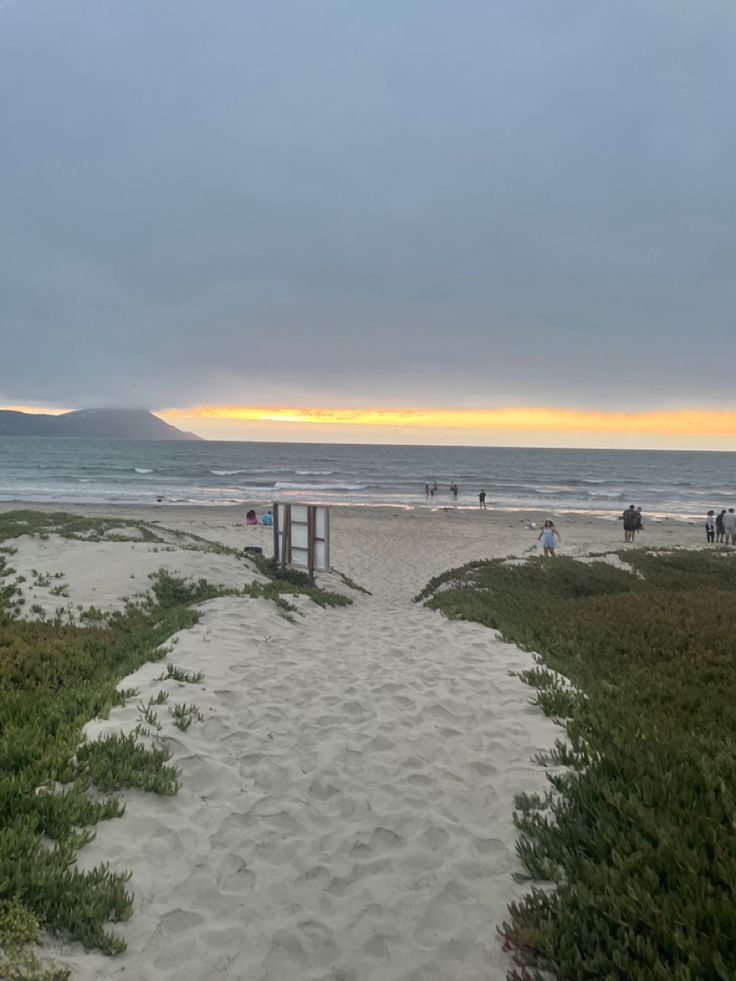 a path leading to the beach with people walking on it and in the distance, there is a body of water