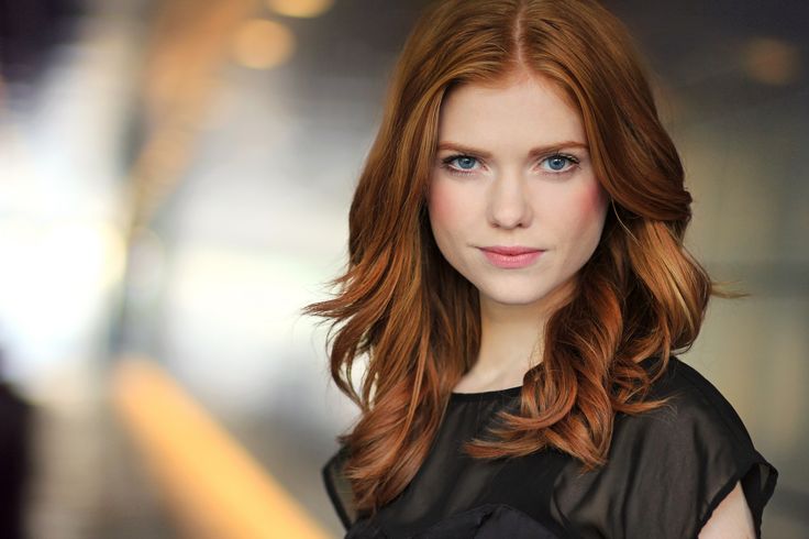 a woman with red hair and blue eyes posing for a photo in front of a wall