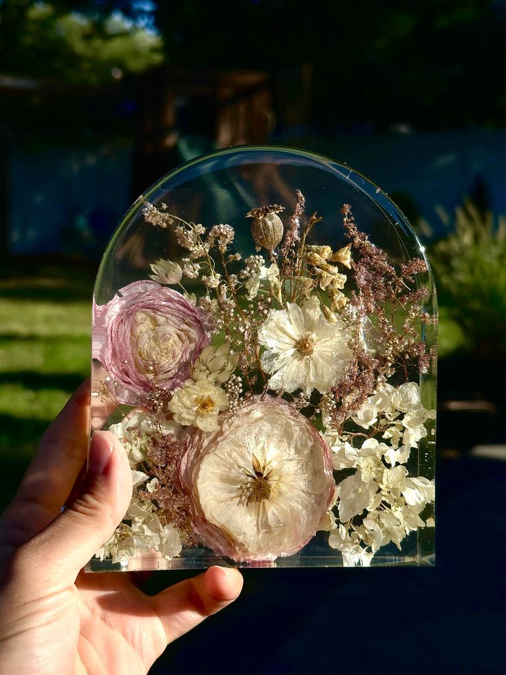 a person holding up a clear glass vase with flowers in it on the grass outside