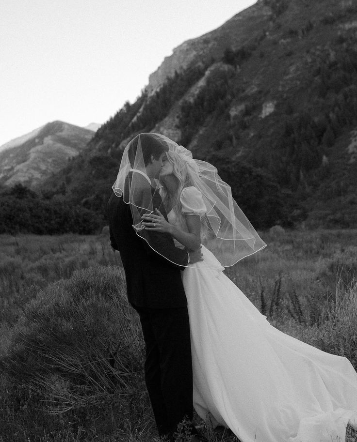 a bride and groom kissing in front of a mountain range with their veil blowing in the wind