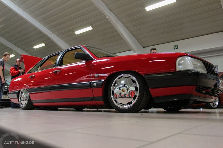 a red car parked in a garage with people standing around and looking at the cars