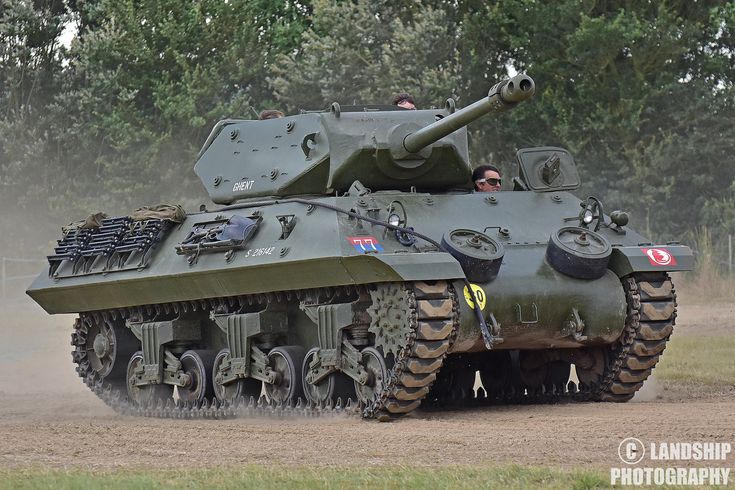 an army tank driving down a dirt road in front of some trees and green grass