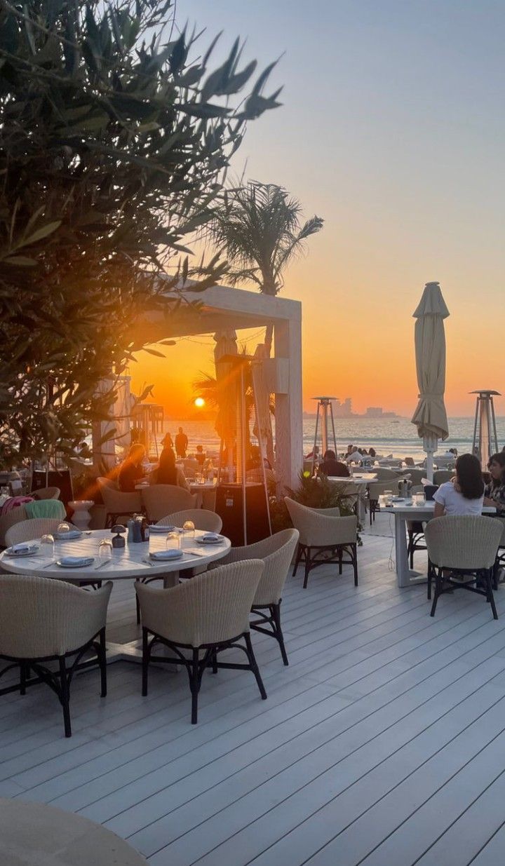 people are sitting at tables on the deck near the ocean as the sun goes down