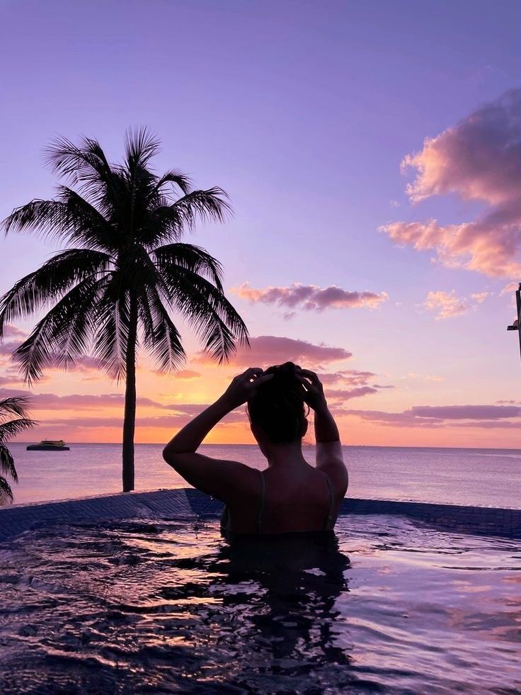 a person sitting in a swimming pool with palm trees and the sun setting behind them