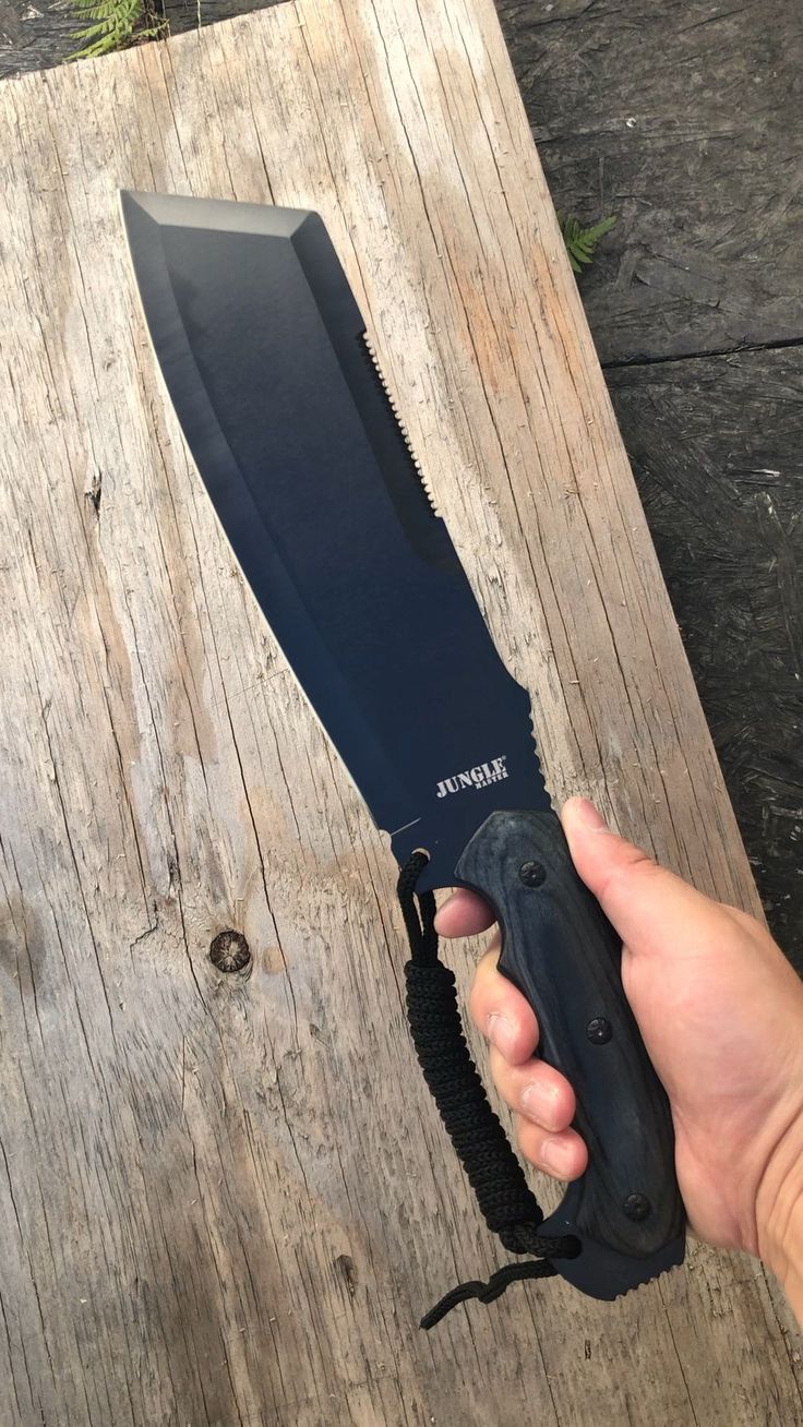 a hand holding a black knife on top of a wooden table next to a piece of wood