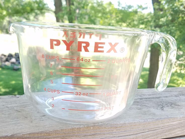 a glass measuring cup sitting on top of a wooden table in front of some trees