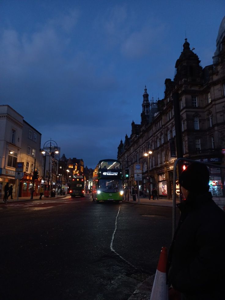 a green double decker bus driving down a city street at night with buildings in the background