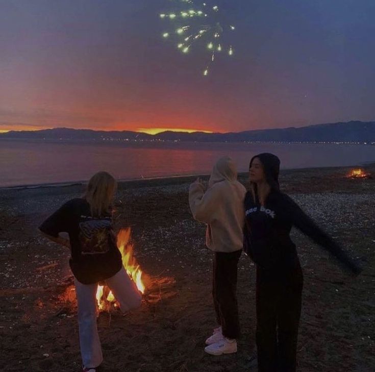 two women standing next to a fire with fireworks in the sky above them at night