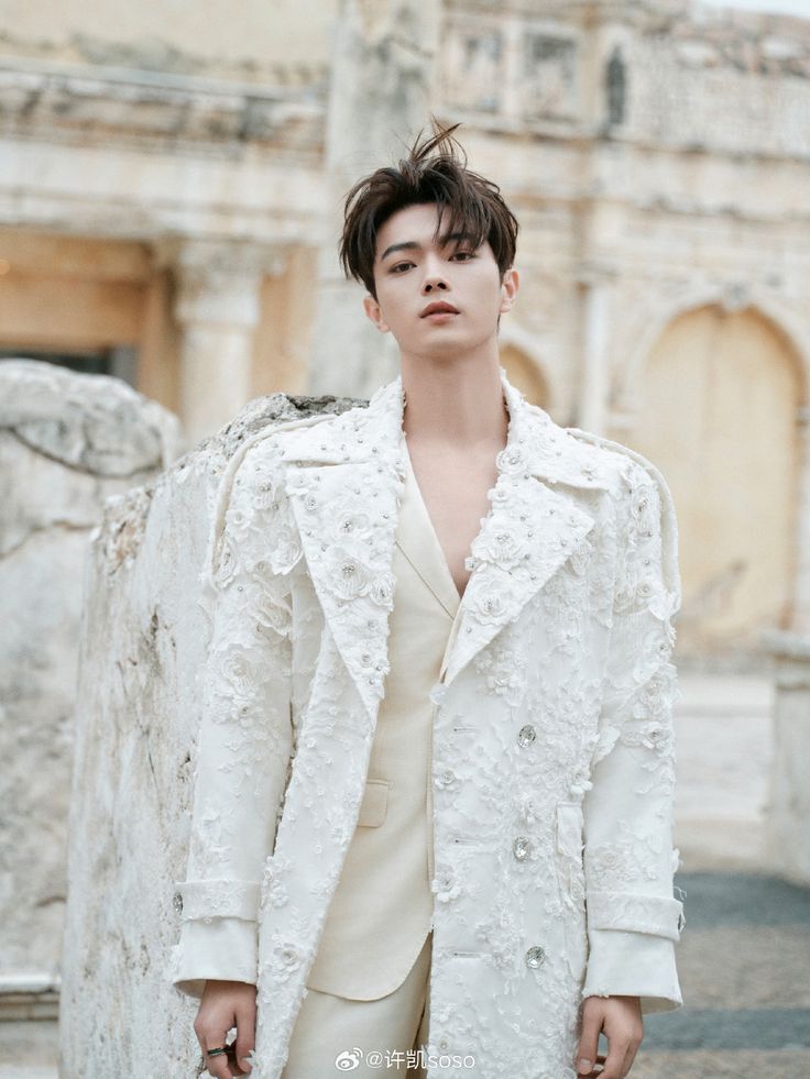 a man in a white coat standing next to a stone wall and looking off into the distance