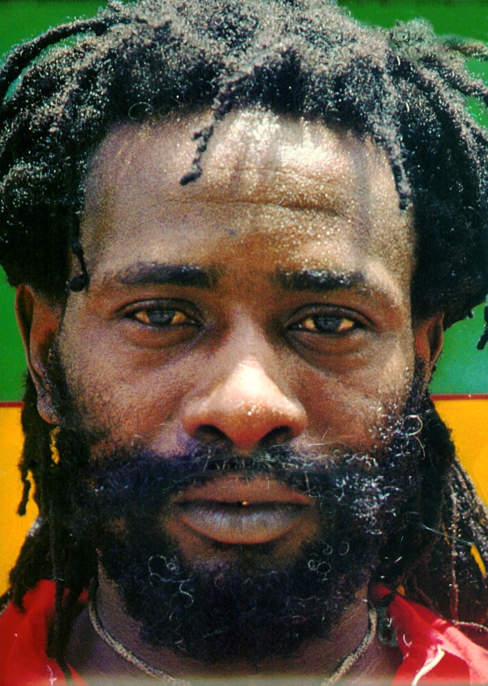 a man with dreadlocks and a red shirt is looking at the camera in front of a multi - colored background