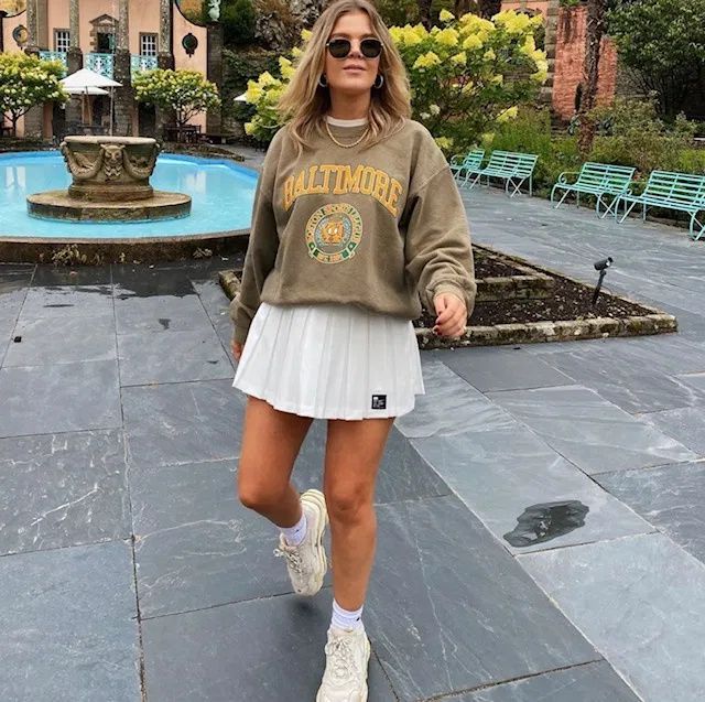 a woman walking down the street in front of a fountain wearing a sweatshirt and pleated skirt