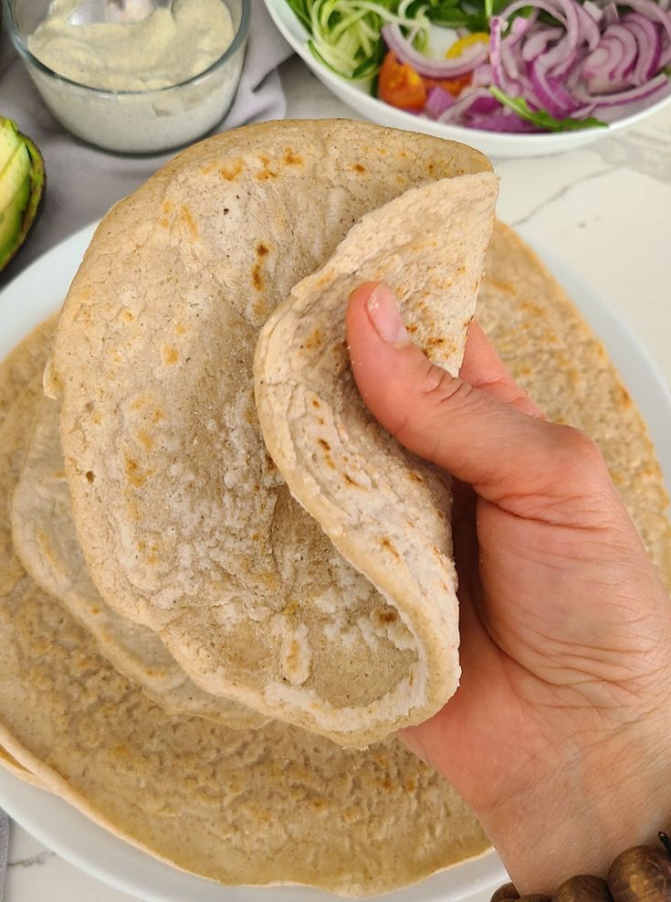 a hand holding up a tortilla on top of a white plate next to bowls of vegetables