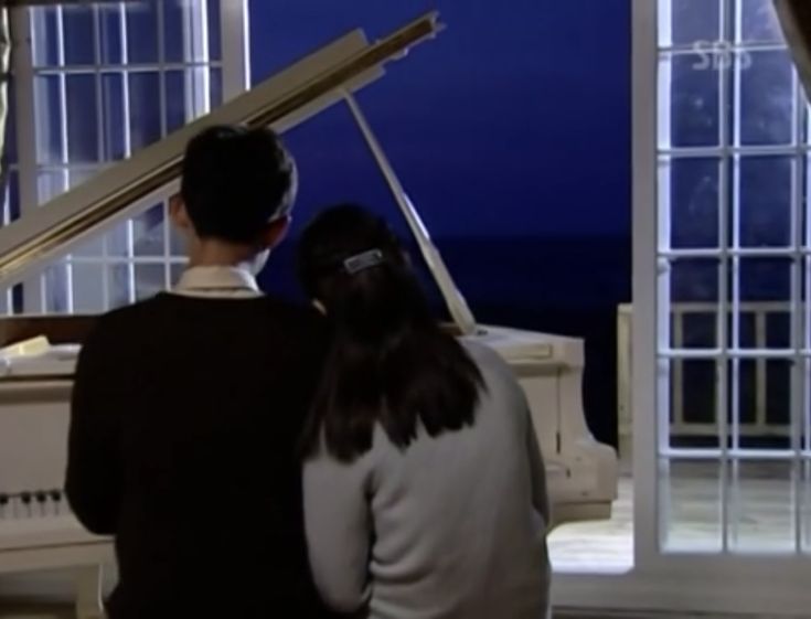 two people sitting at a piano in front of a window looking out onto the ocean