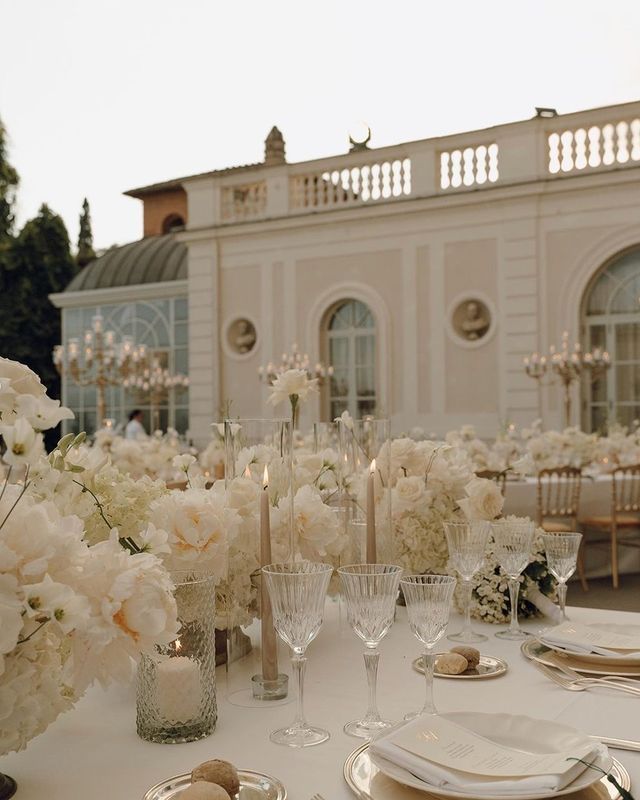 the table is set with white flowers and place settings for an elegant wedding reception in front of a large building