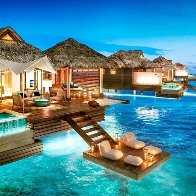 an outdoor swimming pool with lounge chairs and thatched huts in the background at night