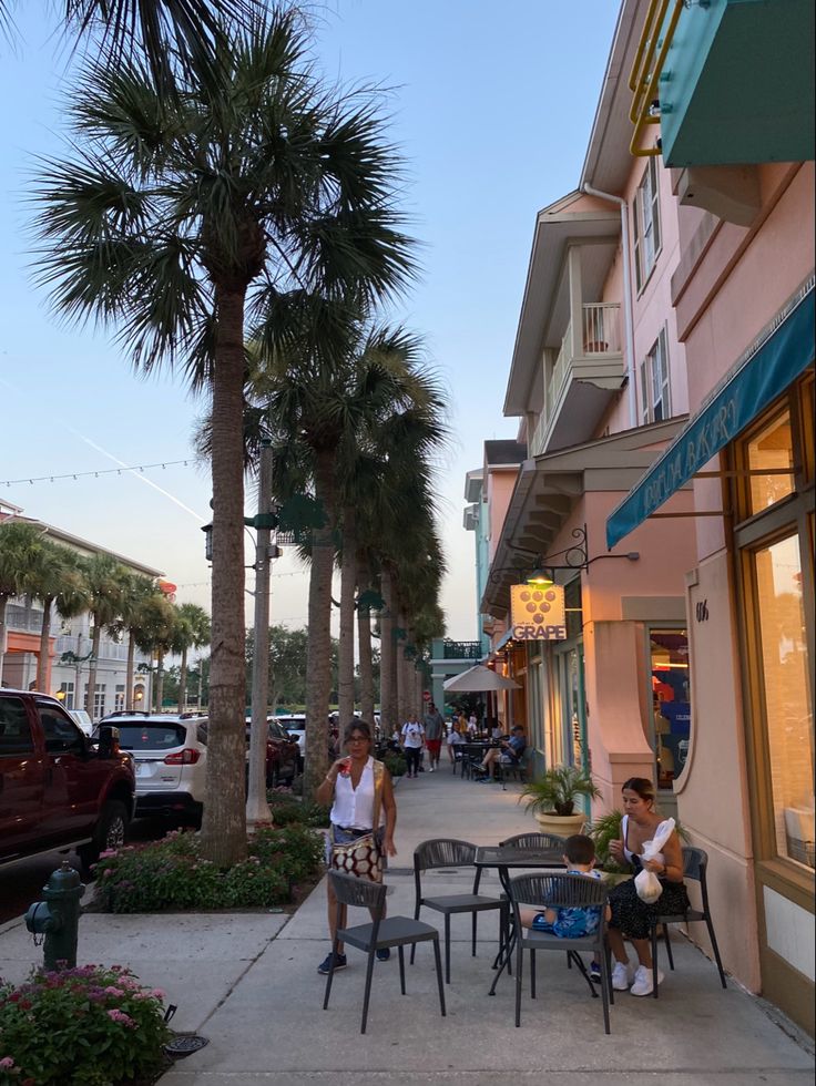 two people sitting at tables on the sidewalk in front of some buildings and palm trees