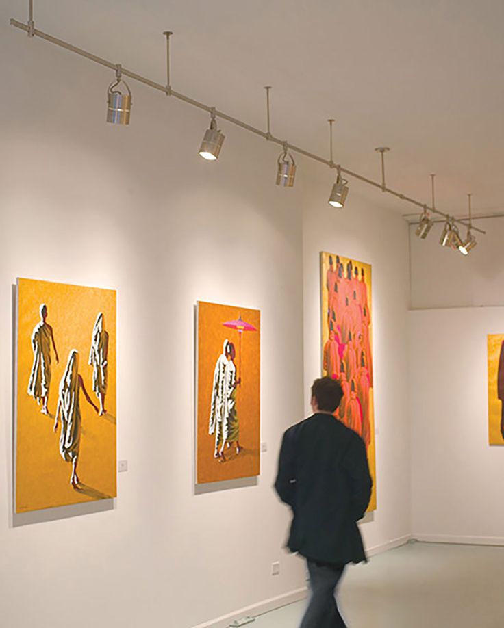 a man walking past paintings on display in an art gallery with lights hanging from the ceiling