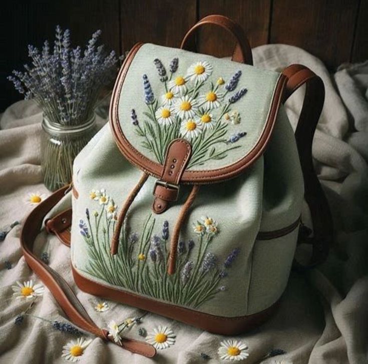 a backpack with flowers painted on it sitting on a blanket next to a vase filled with wildflowers