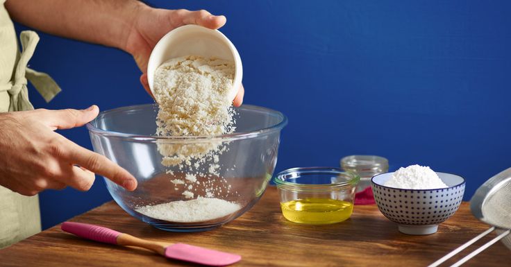 a person pouring flour into a bowl on top of a wooden table next to bowls and measuring spoons