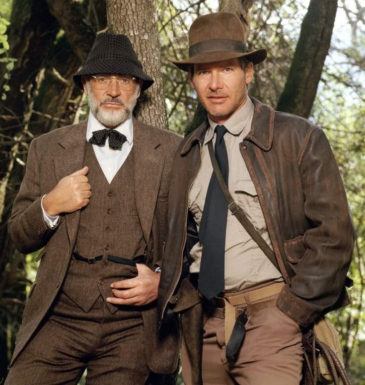 two men in brown suits and hats standing next to each other near a tree trunk