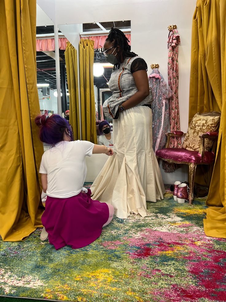 a woman is helping another woman put on her dress in front of a mirror with yellow drapes