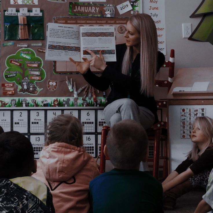 a woman standing in front of a classroom full of children sitting on the floor and reading to them