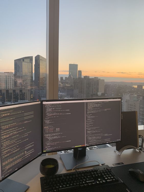 two computer monitors sitting on top of a desk in front of a window with cityscape