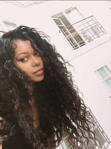 a woman with long curly hair standing in front of a white building and looking at the camera