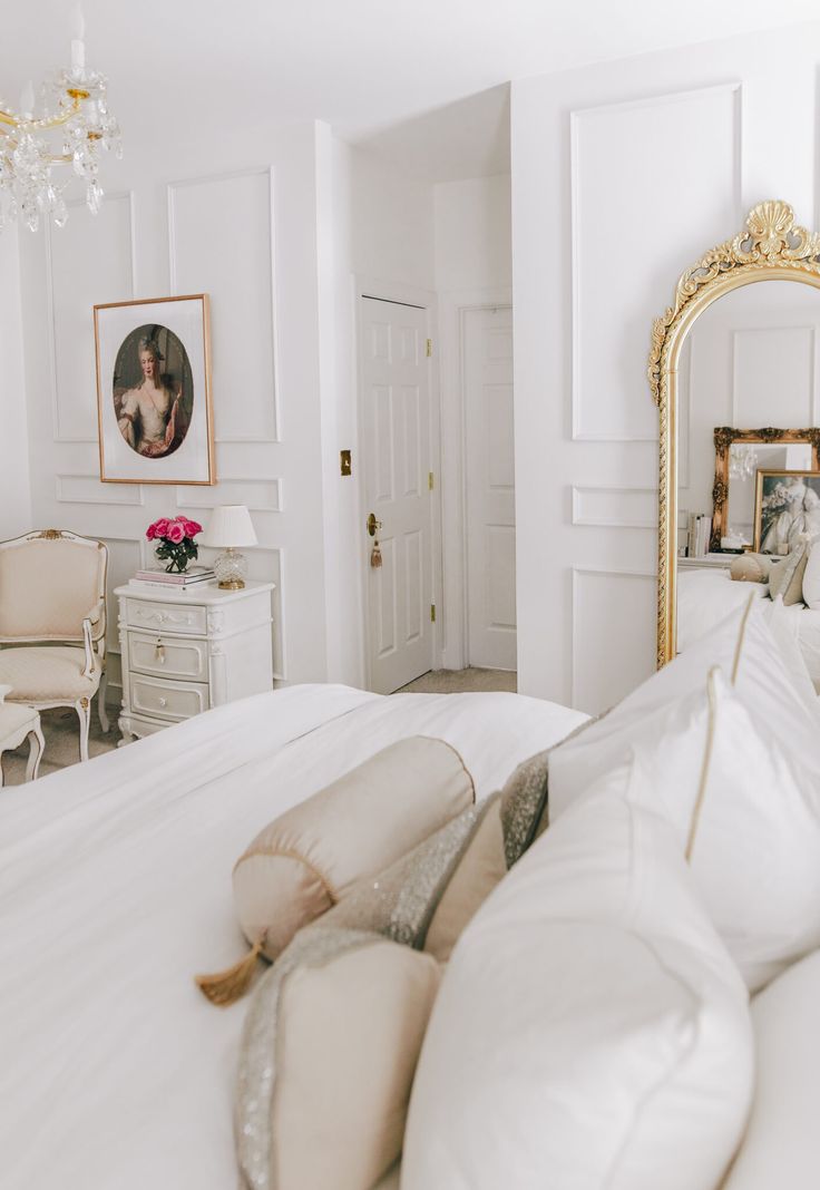 a white bed with pillows and a mirror on the wall next to it in a bedroom