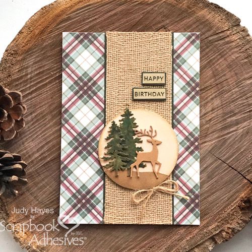 a christmas card with a deer and pine tree on it, sitting on top of a piece of wood