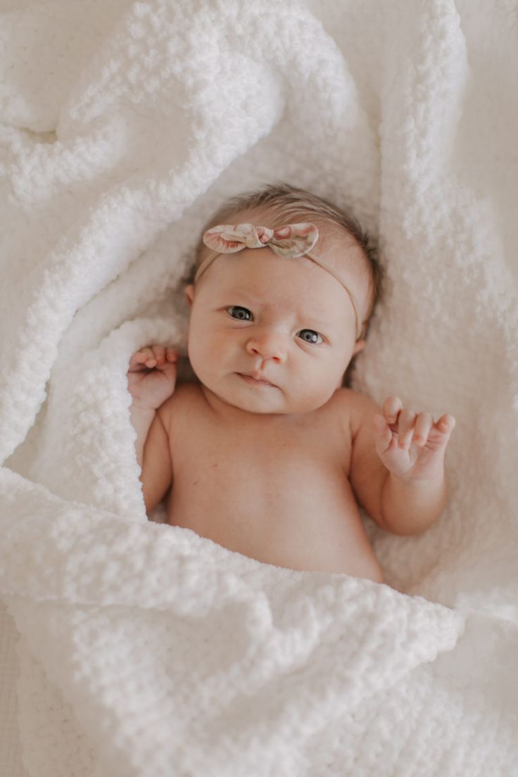 a baby wrapped in a white blanket looking at the camera with one hand on her head