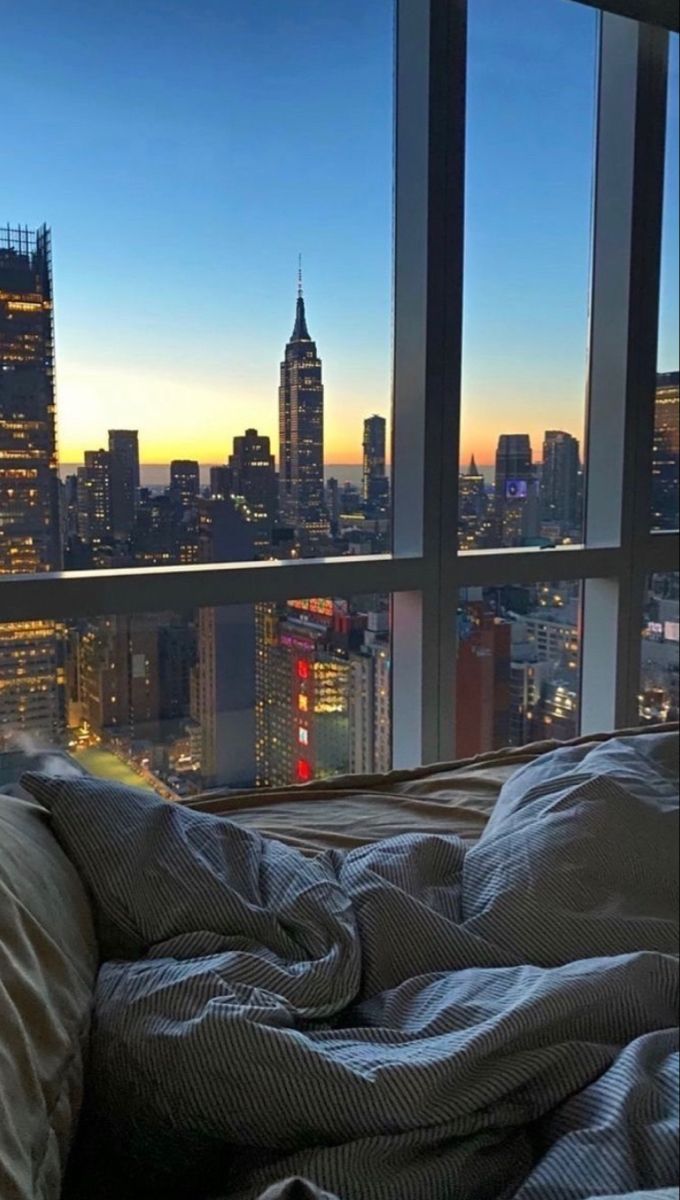 an unmade bed in front of a large window overlooking the city at night time