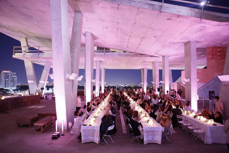 a large group of people sitting at long tables under an awning in the evening