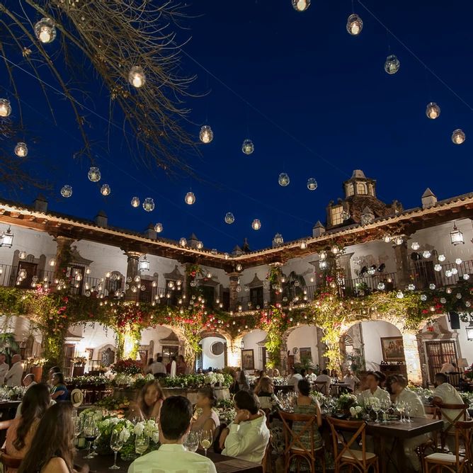 people sitting at tables in an old building with lights hanging from the ceiling and potted plants