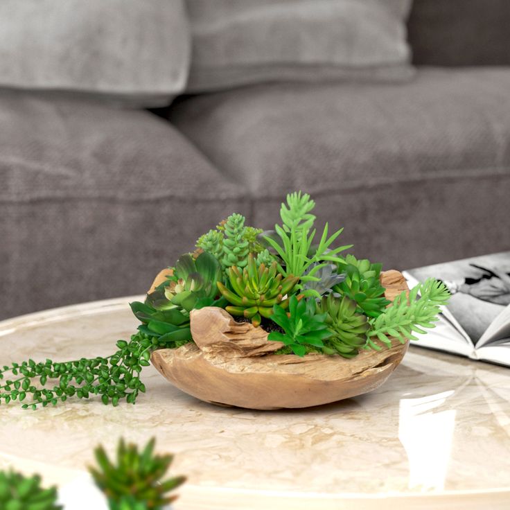 a table topped with a wooden bowl filled with green plants next to a gray couch