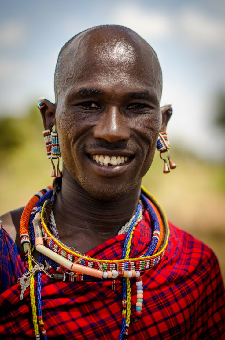 an african man wearing colorful necklaces and headdress smiles at the camera while standing in a field