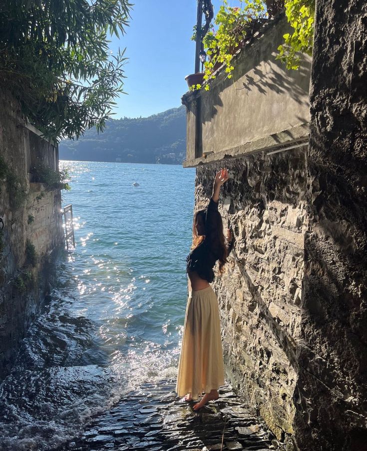 a woman standing on the edge of a stone wall next to water with her hand up in the air