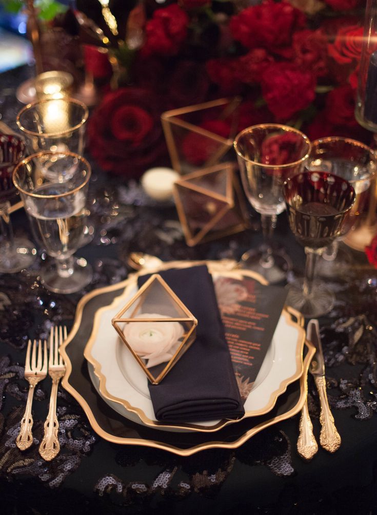 a black and gold table setting with silverware, napkins and red roses in the background